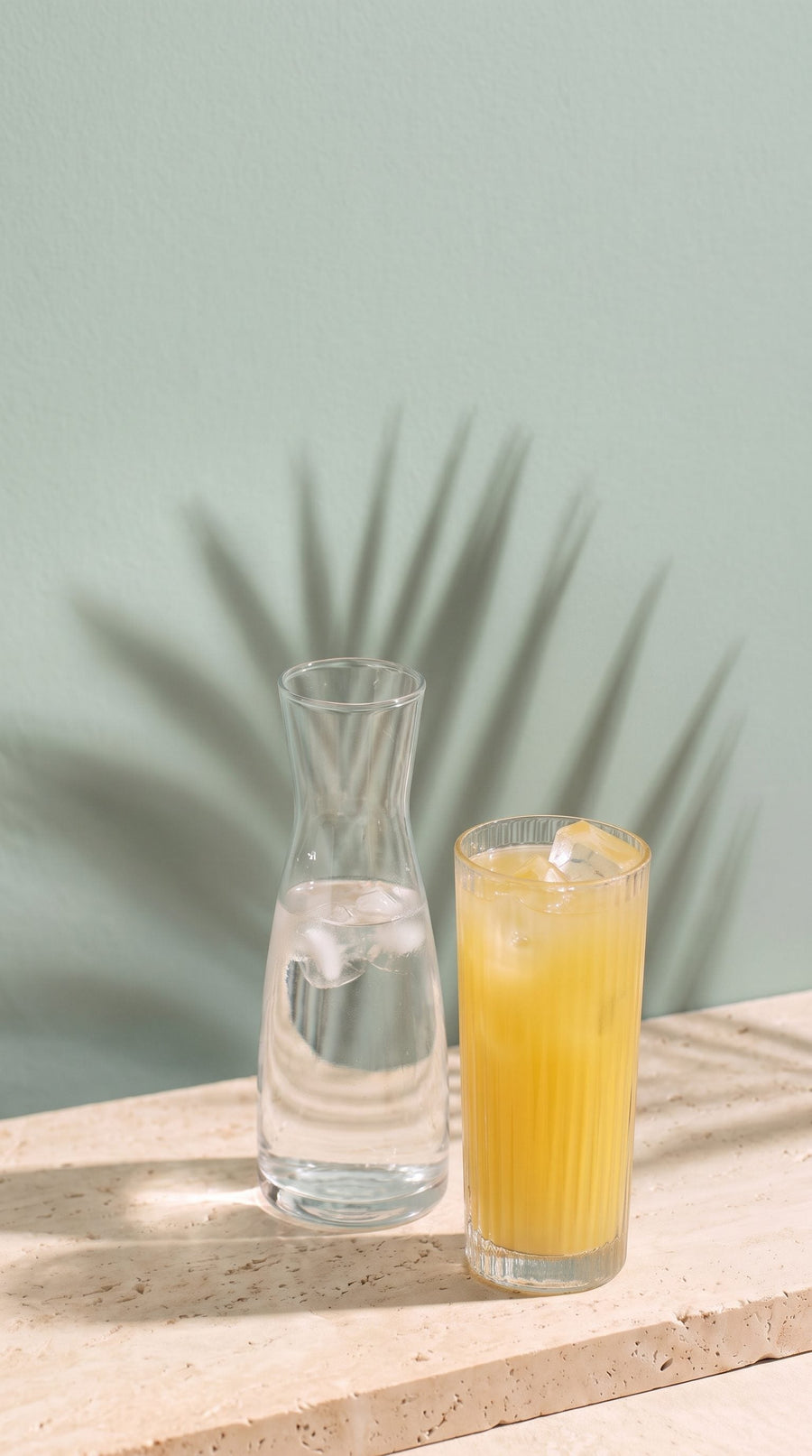 A carafe of iced water beside a glass of golden drink on a travertine ledge in morning sun