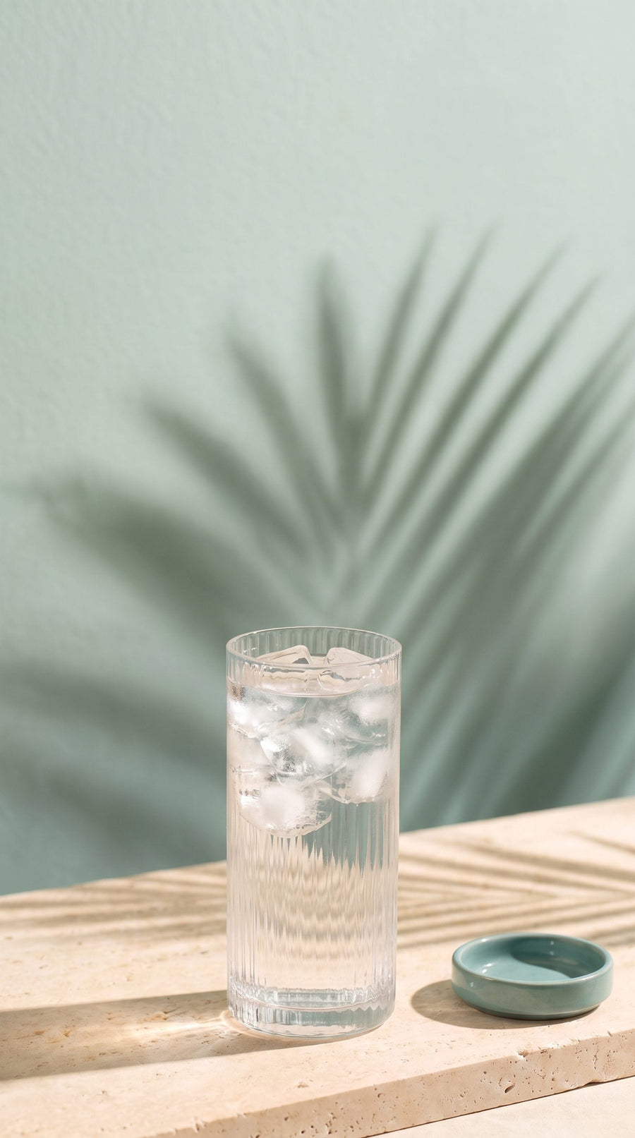 A glass of iced water and an empty teal dish on a stone ledge in early morning light