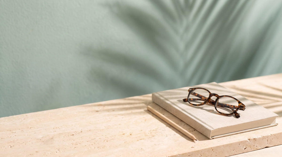 A closed linen notebook, a wooden pencil and folded reading glasses on a travertine ledge in morning sun