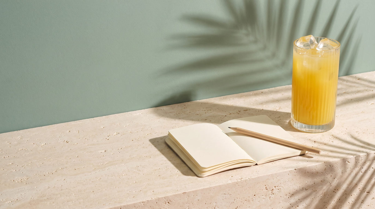 An open blank notebook and pencil beside a glass of golden drink on a stone ledge