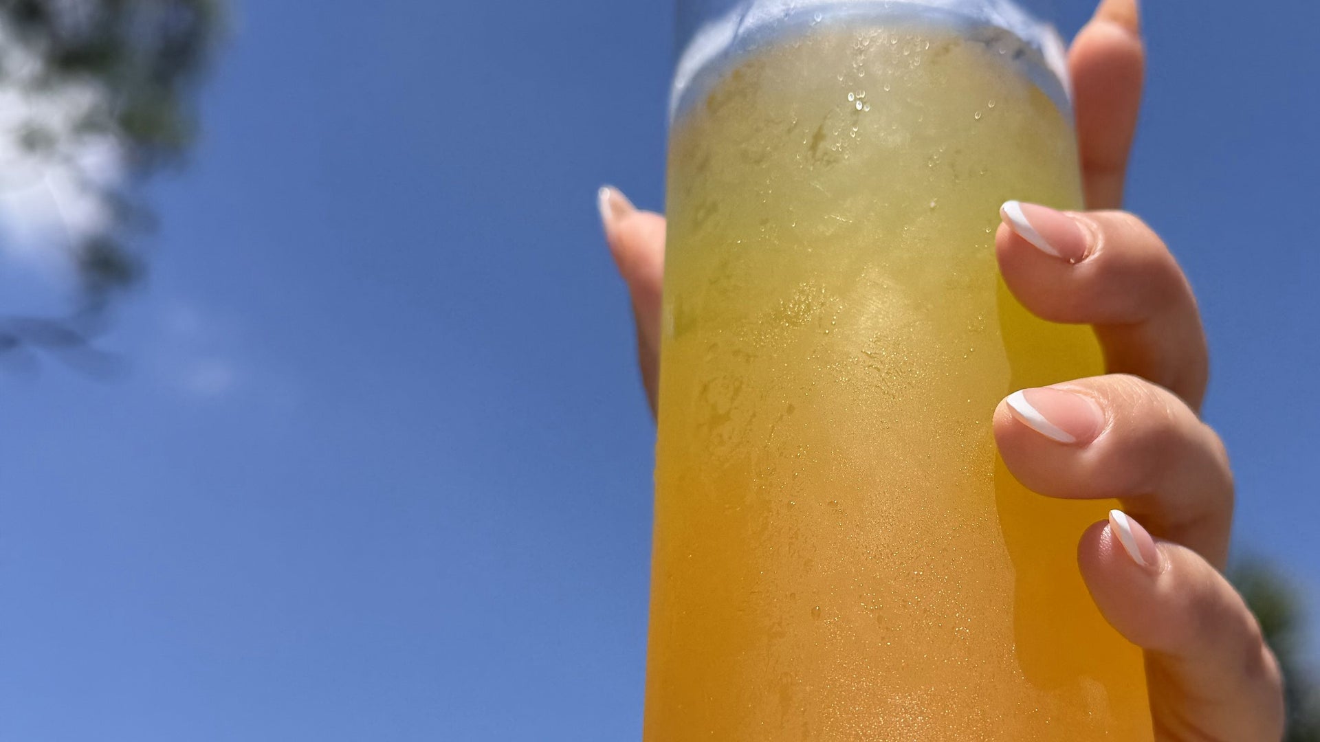 A hand raising a bottle of cloudy golden GLPrelief drink to a blue sky between palms