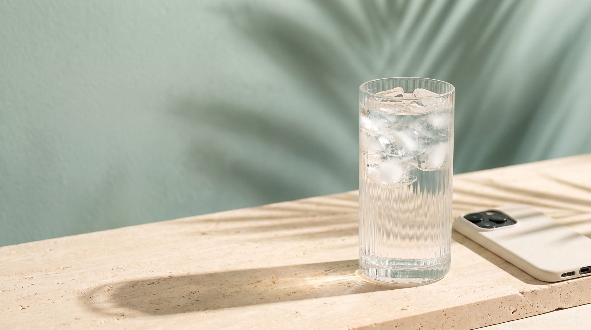 A glass of iced water and a phone lying face-down on a stone ledge in morning sun